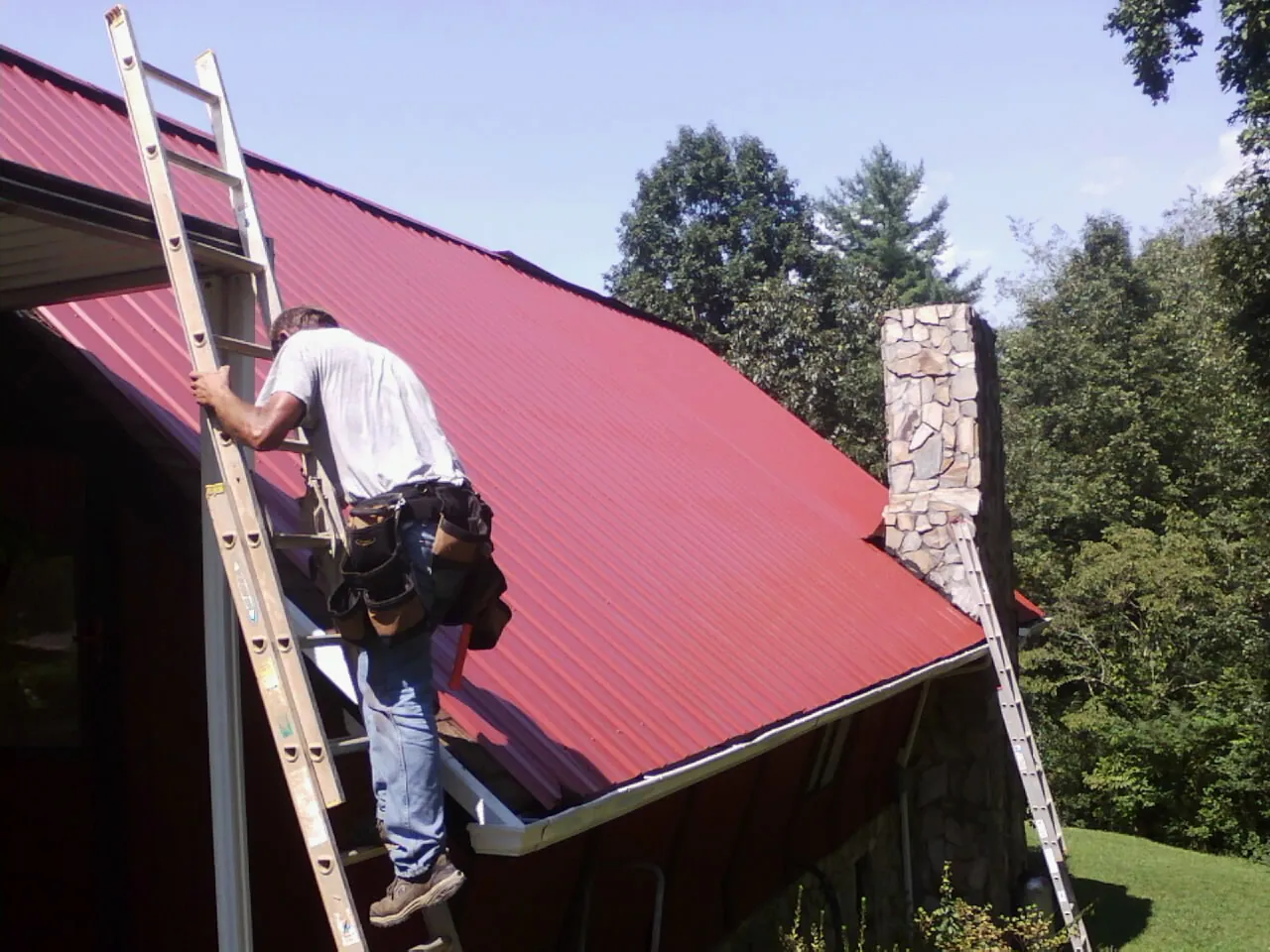 Roofer on a ladder beside a red metal roof and stone chimney — Soffit & Fascia Repair by Roofing Brothers in Ocoee